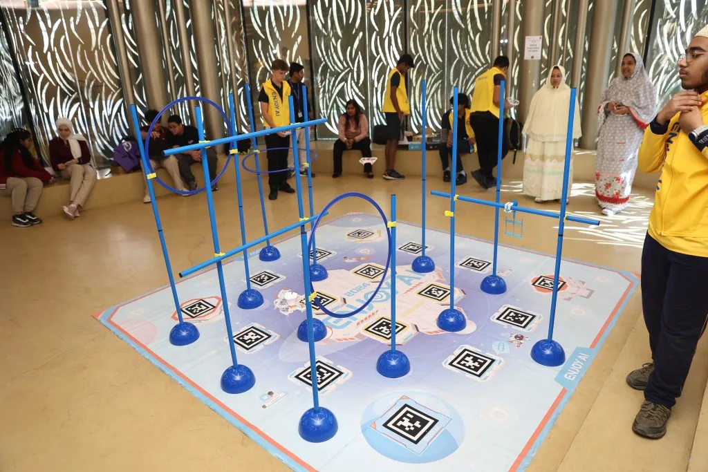 Students in yellow vests around a floor course of blue poles, hoops and printed markers set up for a drone activity, with other students seated nearby