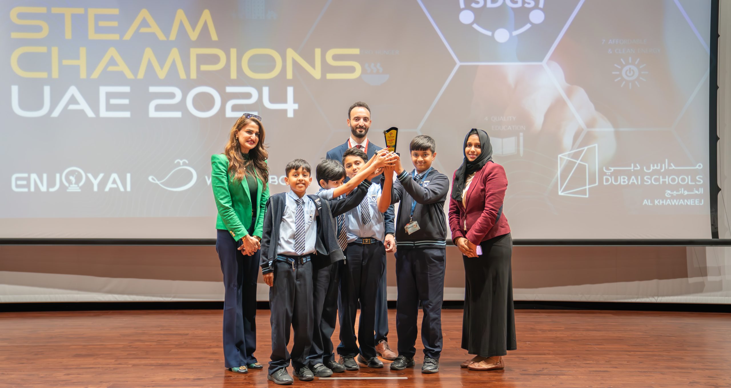 A winning student team and their coaches holding a trophy on stage at STEAM Champions UAE 2024
