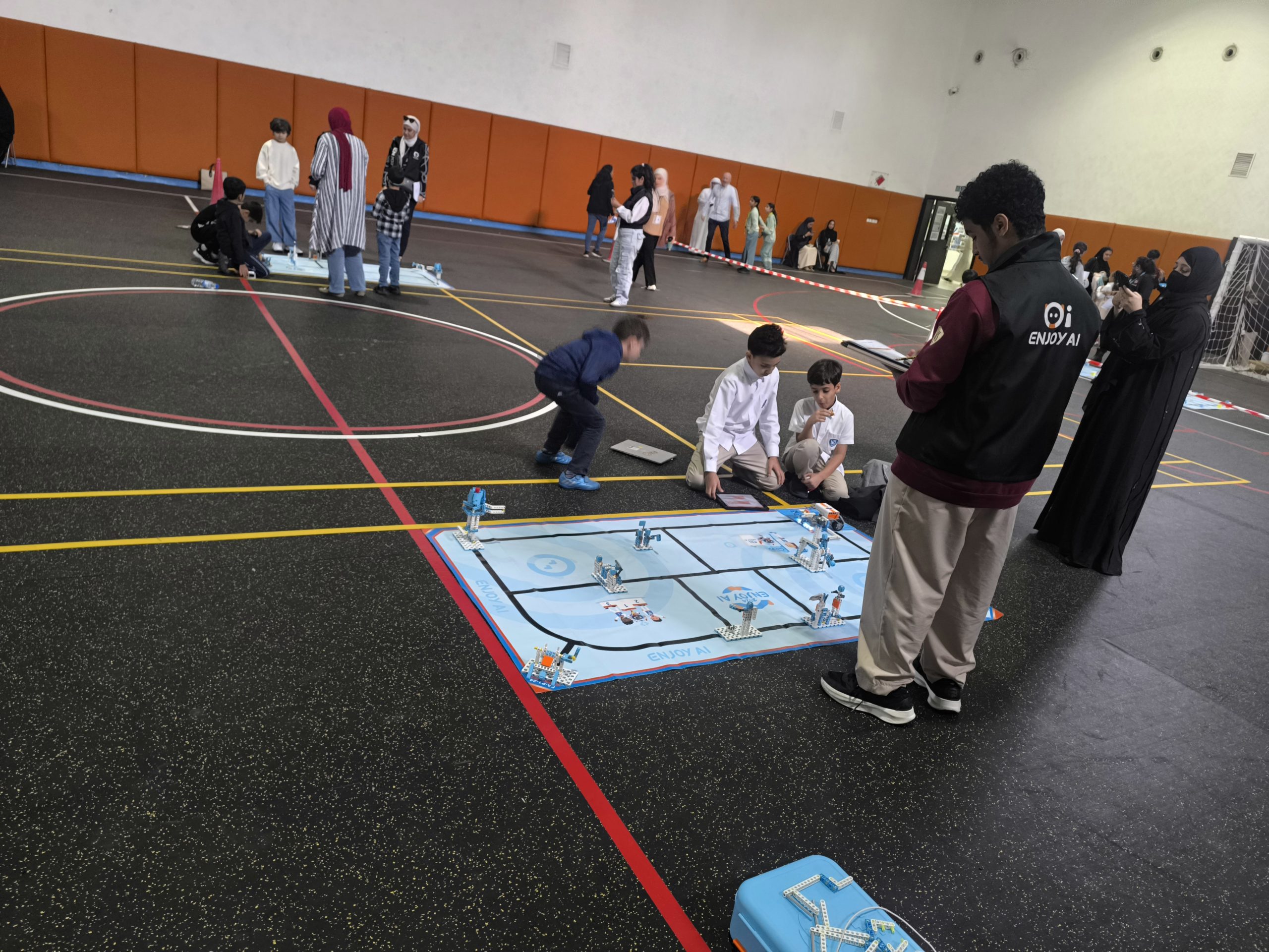 Students competing with robots on a challenge mat while a judge observes in a sports hall
