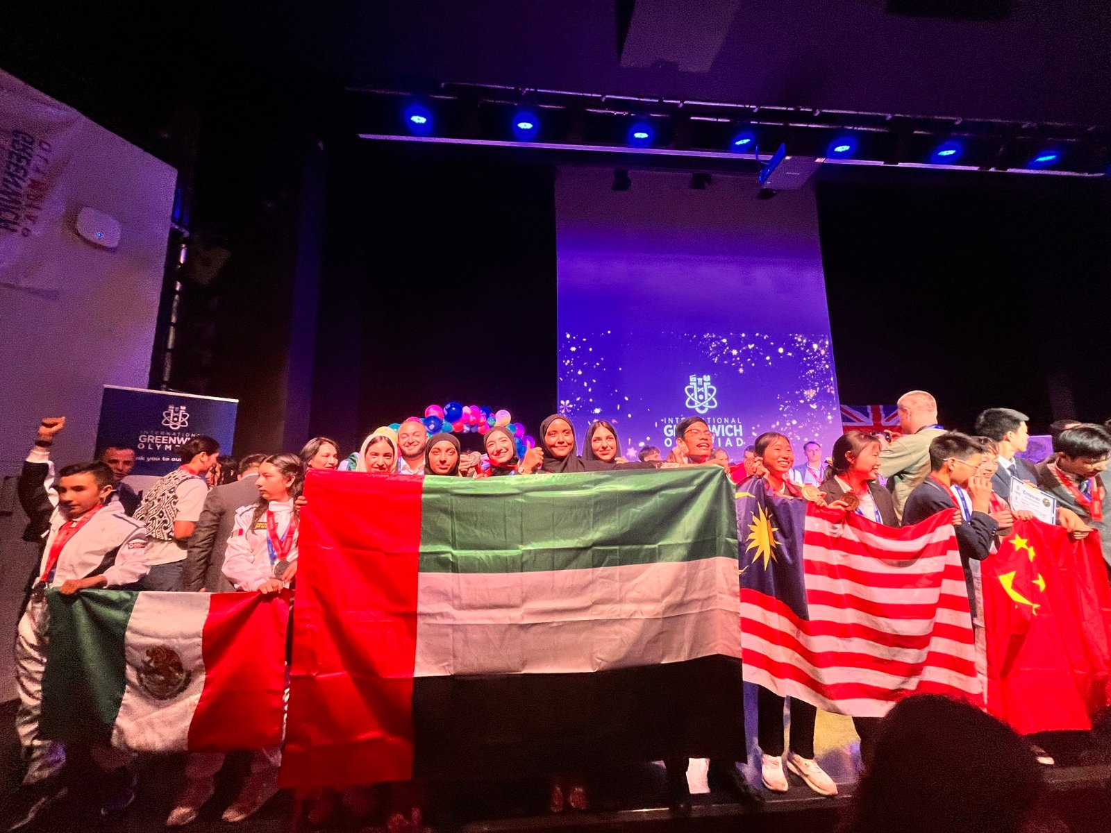 Student teams on stage at an international STEAM Champions olympiad, holding the UAE flag alongside flags of other participating countries