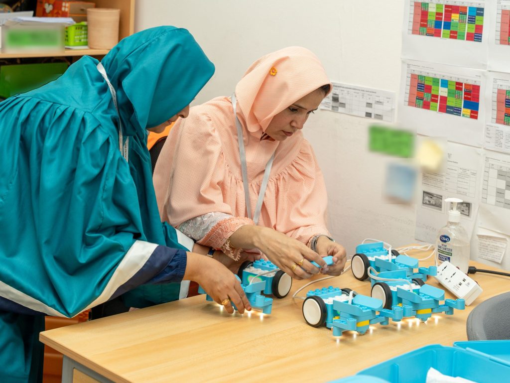 Two women setting up blue wheeled robotics kits on a classroom desk