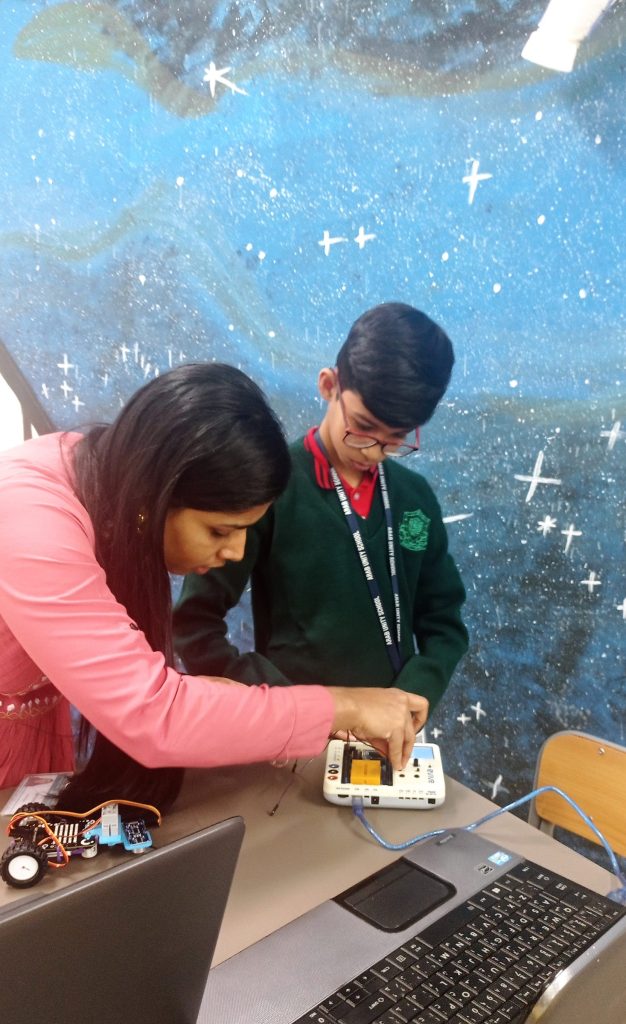 An adult helping a student connect wires to an electronics controller board, with a laptop and a small wheeled robot on the table