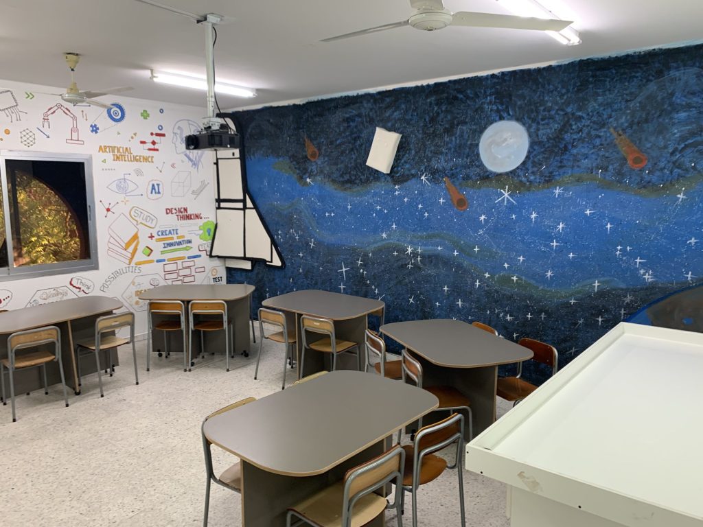 A school classroom with grouped grey tables and chairs, a wall mural of artificial intelligence and design-thinking sketches, a painted space-themed wall and a ceiling-mounted projector