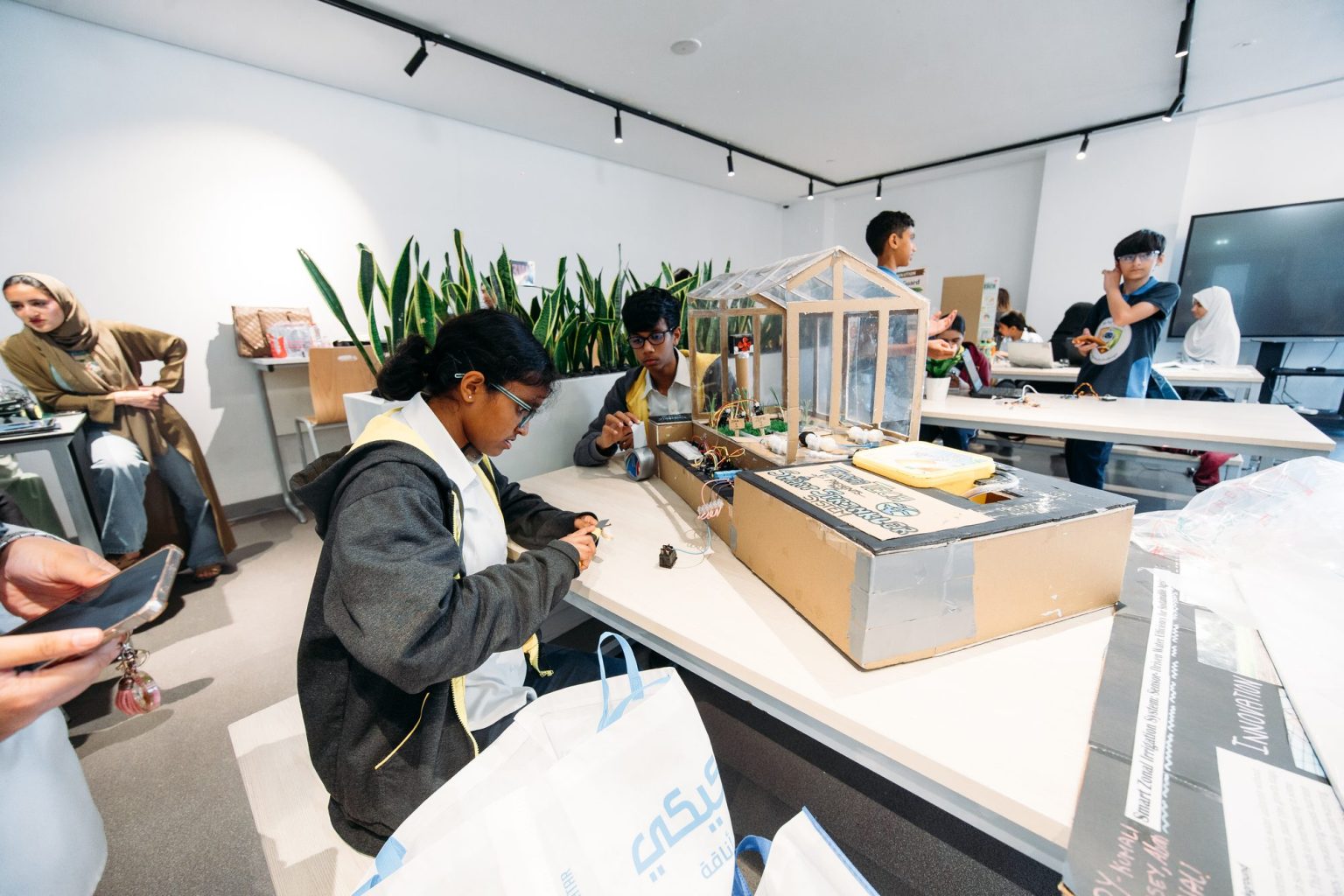A student wiring components beside a cardboard and clear-plastic greenhouse project model, with other students working at tables