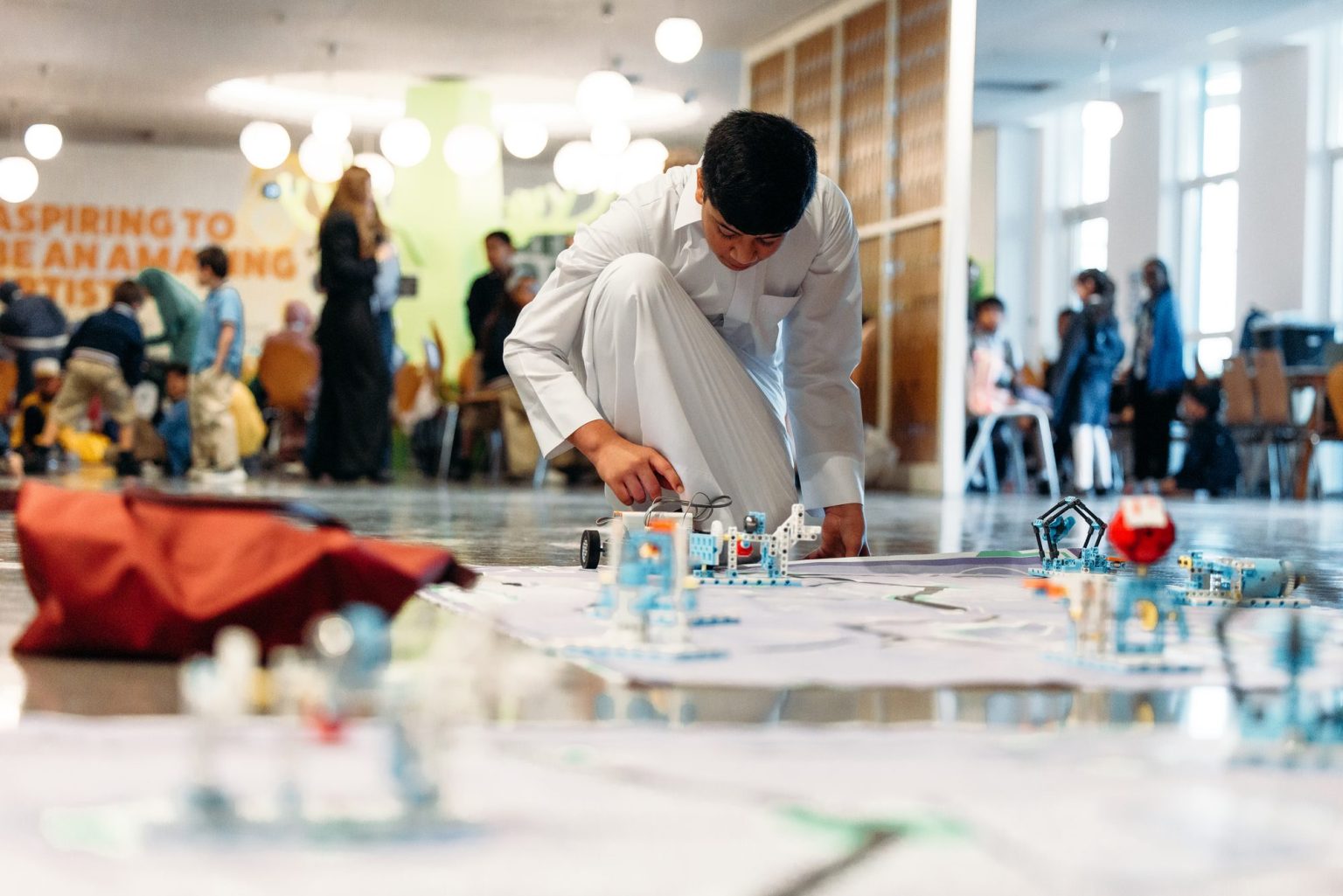 A student kneeling on a competition mat to adjust a robot among robotics models, with other participants in the background