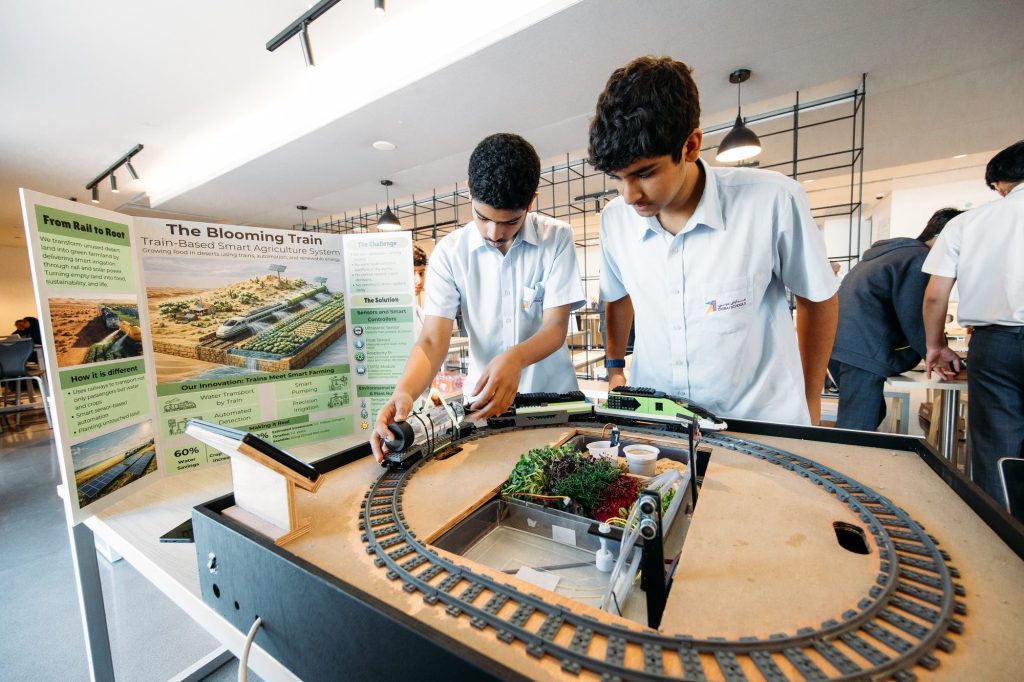 Two students adjusting sensors on a model railway smart-agriculture project with a planted growing tray, beside their project display board
