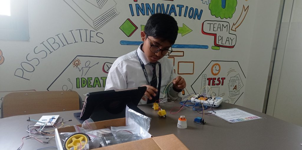 A student connecting jumper wires between small motors, a relay module and a controller board, with an open kit box and a tablet on the table